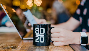 Image shows someone's left hand holding a black mug with the numbers eighty over twenty. There is a laptop in the background with the someone's right hand holding a smartphone