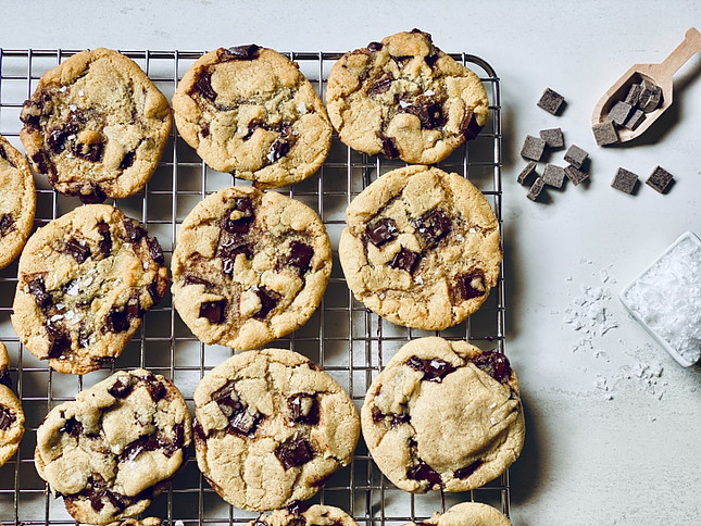 Image shows a grill holding 9 chocolate chip cookies with a wooden spoon holding chocolate chips to the side and a container of salt