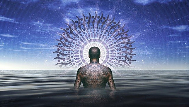 Image of a man with a tatooed back facing away standing waist deep in a body of water with a halo around his head in front of a dark blue sky