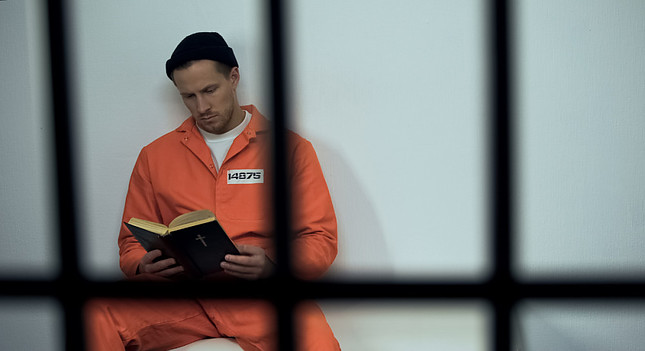 Image shows a prisoner wearing an orange jumpsuit and a beanie while reading a book in the background and a set of prison bars in the foreground