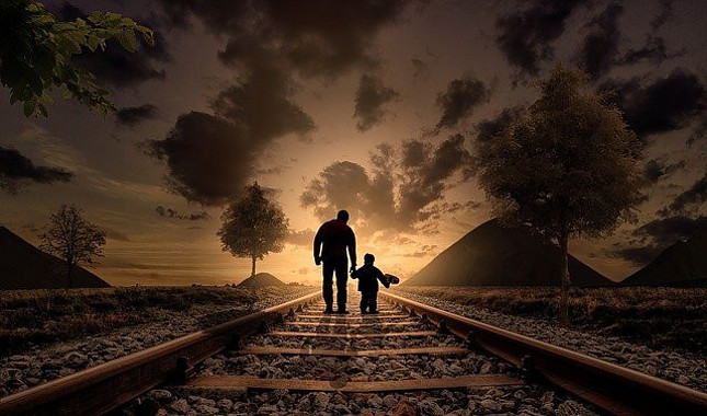 Image shows the silhouette of a grown man holding the hand of a little boy facing away while walking on train tracks