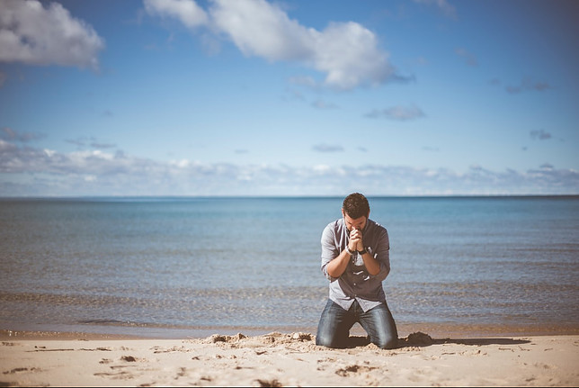 Image shows a man in jeans and a shirt kneeling down on a beach with his head on his hands in prayer