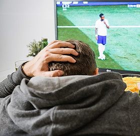 Image shows a man in a hoodie with his hand on his head watching a football player on a television screen