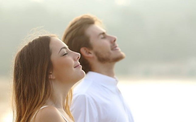 Image shows a woman in the foreground with her eyes closed and her nose up, smiling, in the background is a man with his eyes closed and his nose up, smiling