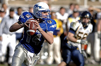 Image shows in the foreground a quarterback in a blue top with silver pants holding a football, ready to pass. The background shows an opposing player and the sidelines.